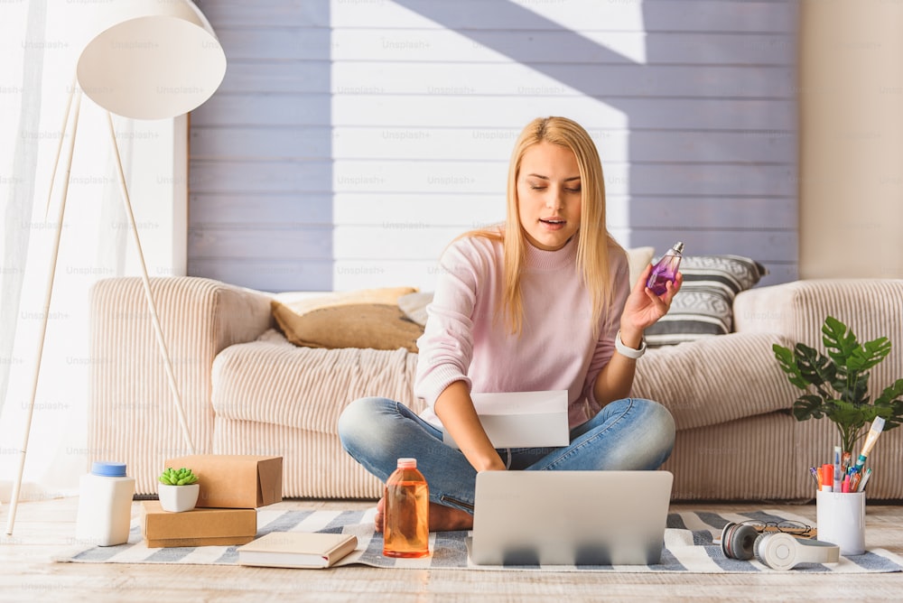 Blond girl is buying cosmetic products in internet with aspiration. She is typing on laptop while holding bottle of perfume. Lady is sitting on floor in her apartment and smiling