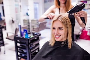見分けのつかないプロの美容師の手が、彼女のクライアント、新しいヘアカット、ブロンドの女性客の髪を乾かしています。