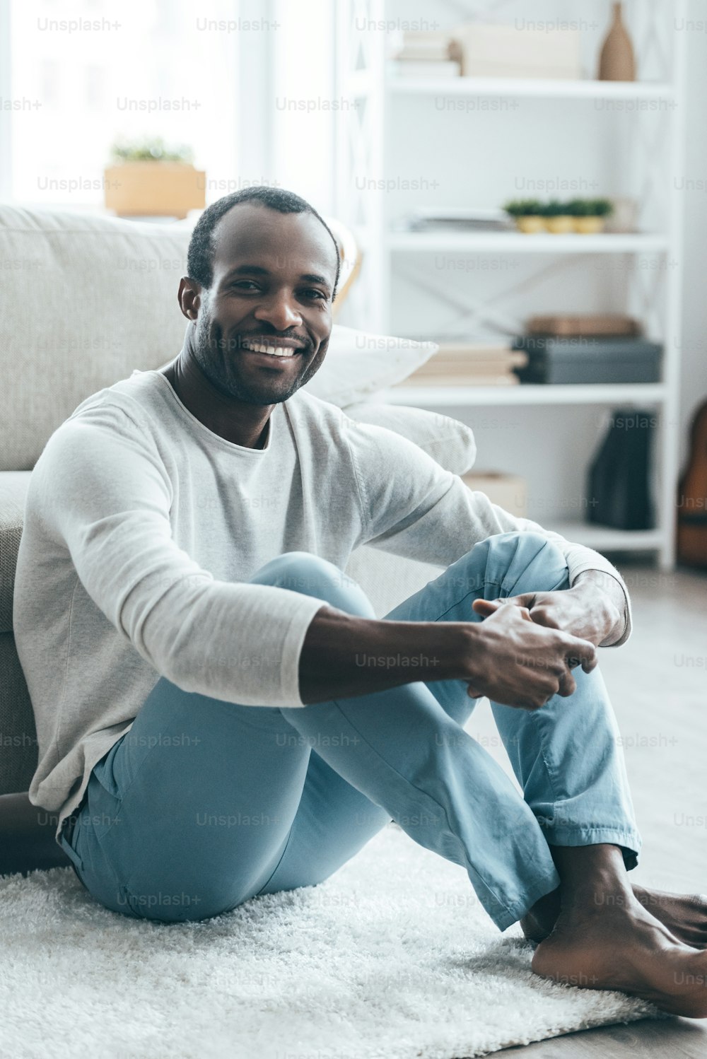 Hübscher junger afrikanischer Mann in eleganter Freizeitkleidung, der in die Kamera schaut und lächelt, während er zu Hause auf dem Teppich sitzt