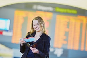 Giovane donna d'affari elegante che tiene il passaporto e la carta d'imbarco, in piedi vicino al bordo delle informazioni di volo nel terminal dell'aeroporto internazionale