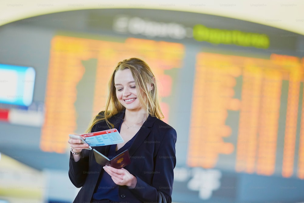 Young elegant business woman holding passport and boarding pass, standing near flight information board in international airport terminal