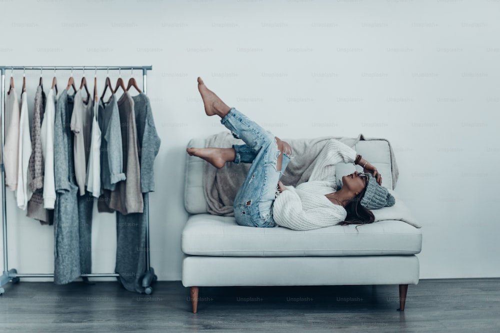 Thoughtful young woman in casual wear touching her head with hand and putting feet up while lying on sofa at home near her clothes hanging on the racks