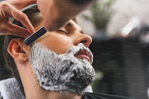 男性のあごひげを剃るためにかみそりを使用する美容師の手の接写。青年はリラックスして目を閉じた