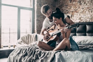Joven guapo barbudo enseñando a su novia a tocar la guitarra mientras ambos están sentados juntos en la cama de su casa