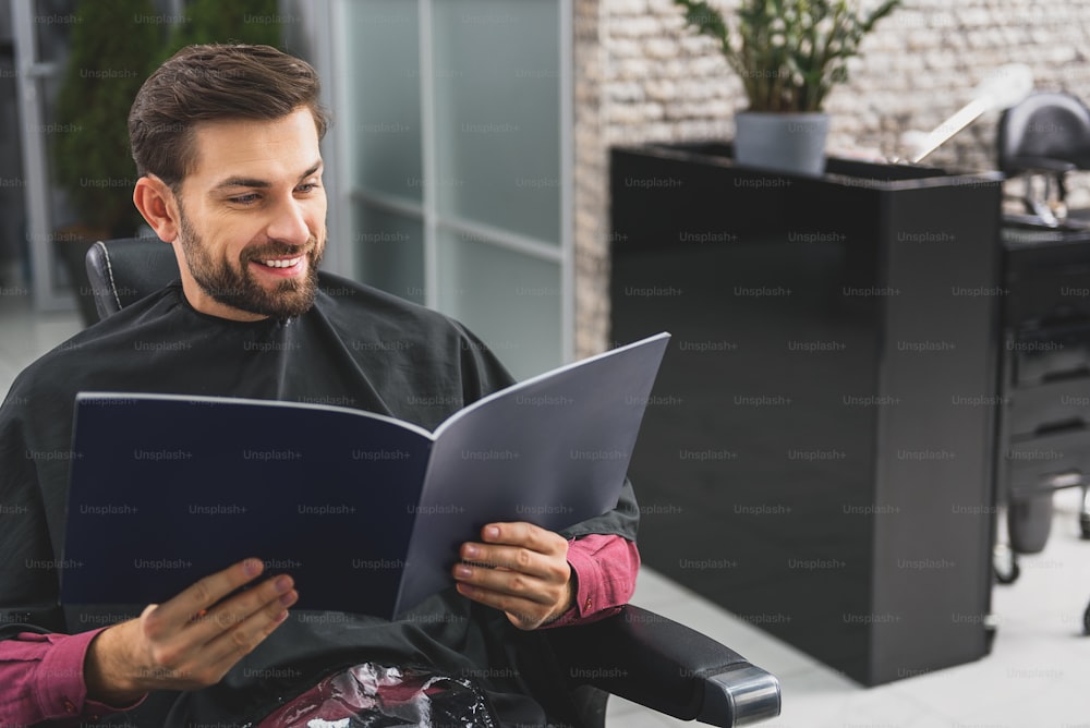 Un joven alegre está leyendo una revista de moda en las peluquerías. Está sentado envuelto en una capa y riendo