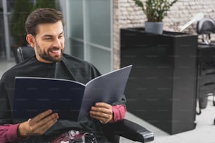 Joyful young man is reading fashion magazine at hairdressers. He is siting wrapped by cape and laughing