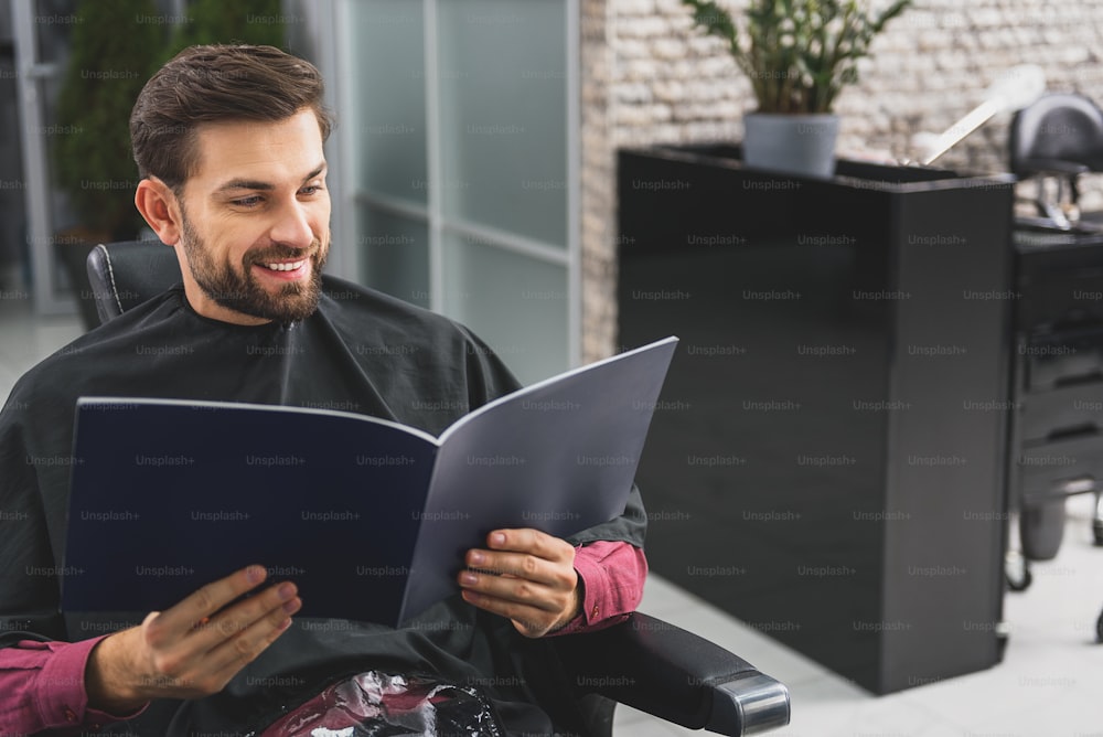 Joyful young man is reading fashion magazine at hairdressers. He is siting wrapped by cape and laughing