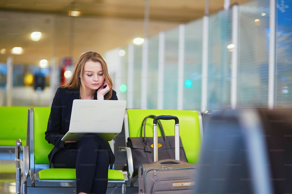 Joven elegante mujer de negocios con equipaje de mano en la terminal del aeropuerto internacional, trabajando en su computadora portátil mientras espera el vuelo