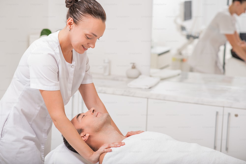 Happy female beautician is doing massage for senior man. She is standing and smiling