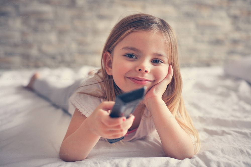 Little girl watching TV lying on bed with remote control in hand.