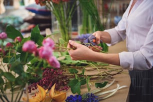 Close up of female arms making bouquet in workshop. Woman is holding scissors near branches of roses