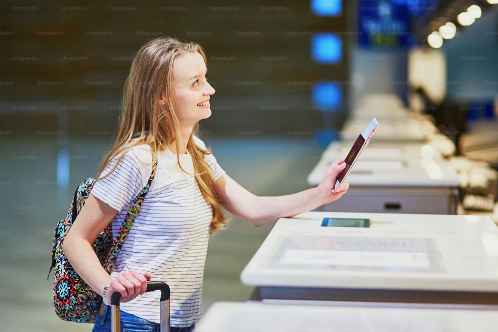 Hermosa joven turista con mochila y equipaje de mano en el aeropuerto internacional en el mostrador de facturación, dando su pasaporte a un oficial y esperando su tarjeta de embarque