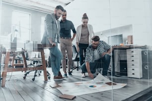 Group of confident business people discussing something while standing in creative office and pointing at large paper lying on floor