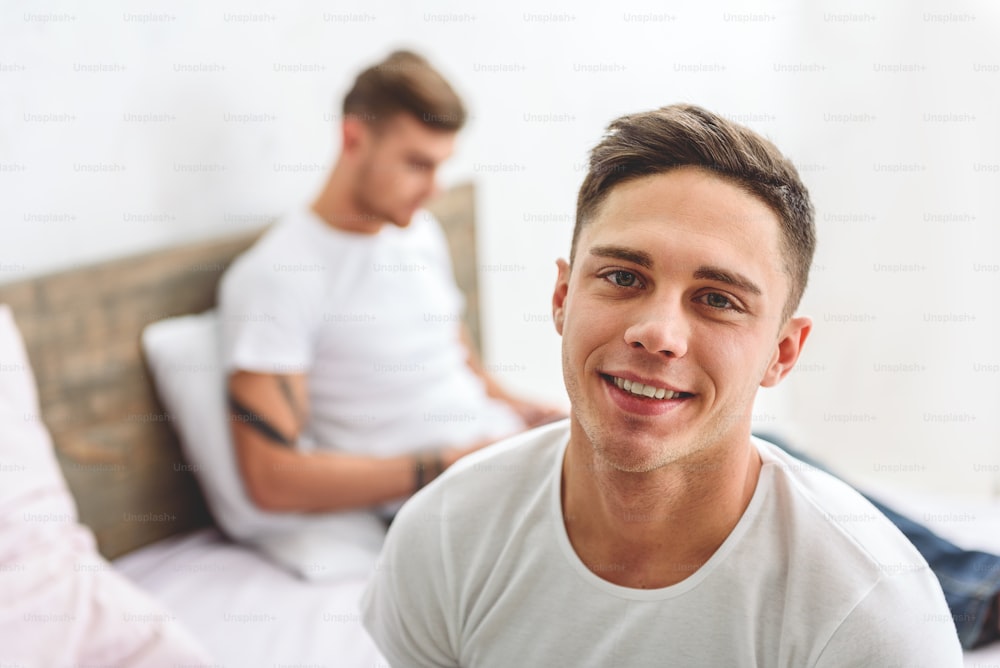 Happy young gay is looking at camera and laughing. His partner is lying on bed and using tablet