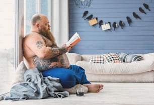 Ruhiger dicker Mann liest Buch mit Konzentration. Er sitzt auf dem Boden und lehnt sich an das Fenster