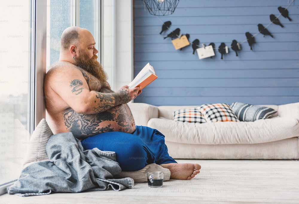 Homem gordo calmo está lendo livro com concentração. Ele está sentado no chão e apoiado na janela.