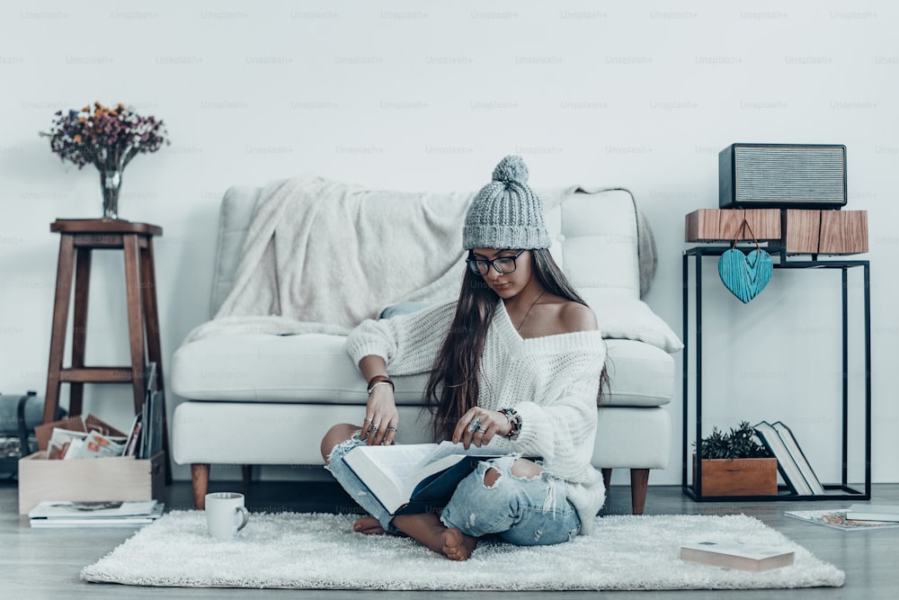 Attractive young woman in casual wear and knit hat reading book while sitting on the floor at home