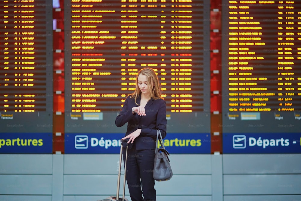 Jovem mulher de negócios elegante com bagagem de mão no terminal do aeroporto internacional, olhando para o quadro de informações, verificando seu voo. Tripulante de cabine com mala.