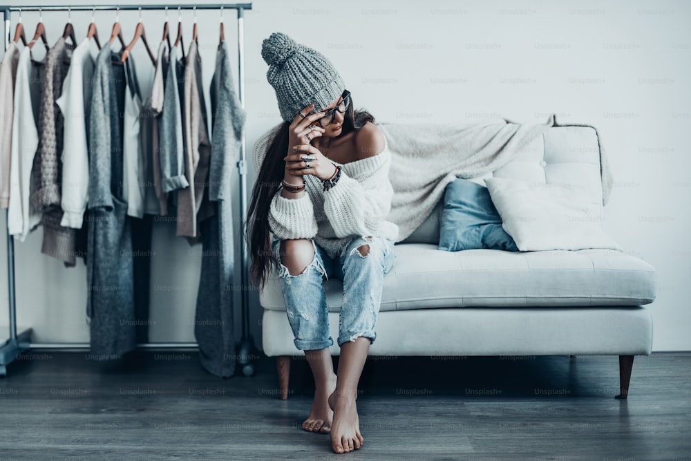Thoughtful young woman in casual wear touching her head with hand looking away while sitting on the couch at home near her clothes hanging on the racks