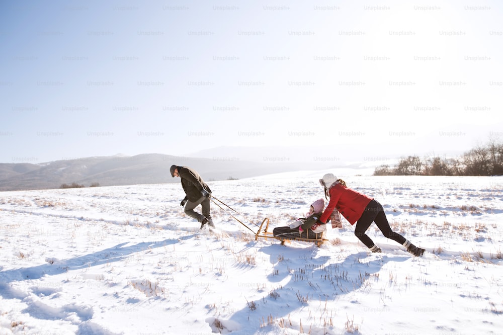 Mother pushing daughter on sledge, father pulling her. Beautiful family in sunny winter nature.