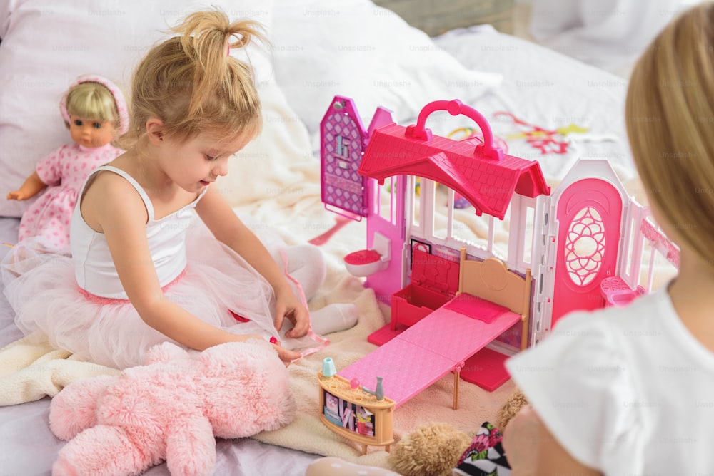 Linda criança pequena está desfrutando de jogo na casa de bonecas com sua irmã mais velha. Ela está sentada na cama e sorrindo