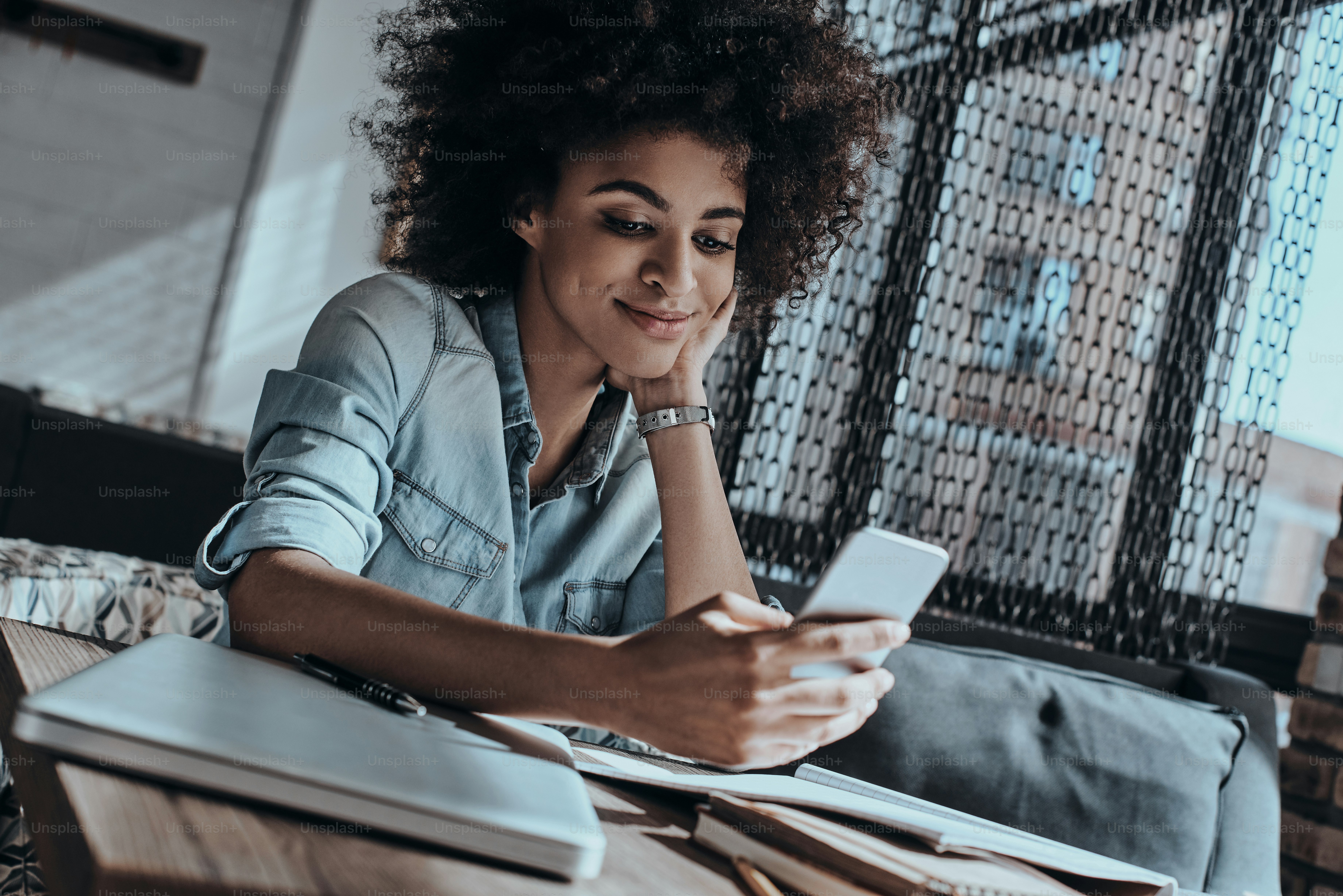 Wanita muda Afrika yang cantik melihat ponsel pintar dan tersenyum sambil duduk di kafe atau kantor kreatif