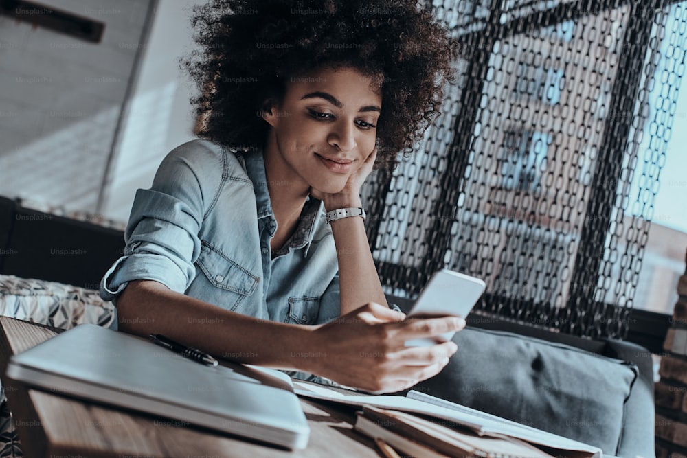 Beautiful young African woman looking at smart phone and smiling while sitting in in cafe or creative office