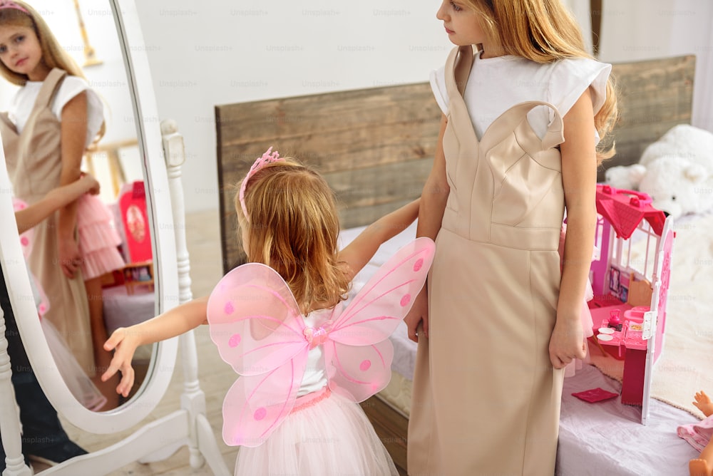 I am the real princess. Excited child is posing near mirror while wearing wings and crown. Her elder sister is trying on dress with interest