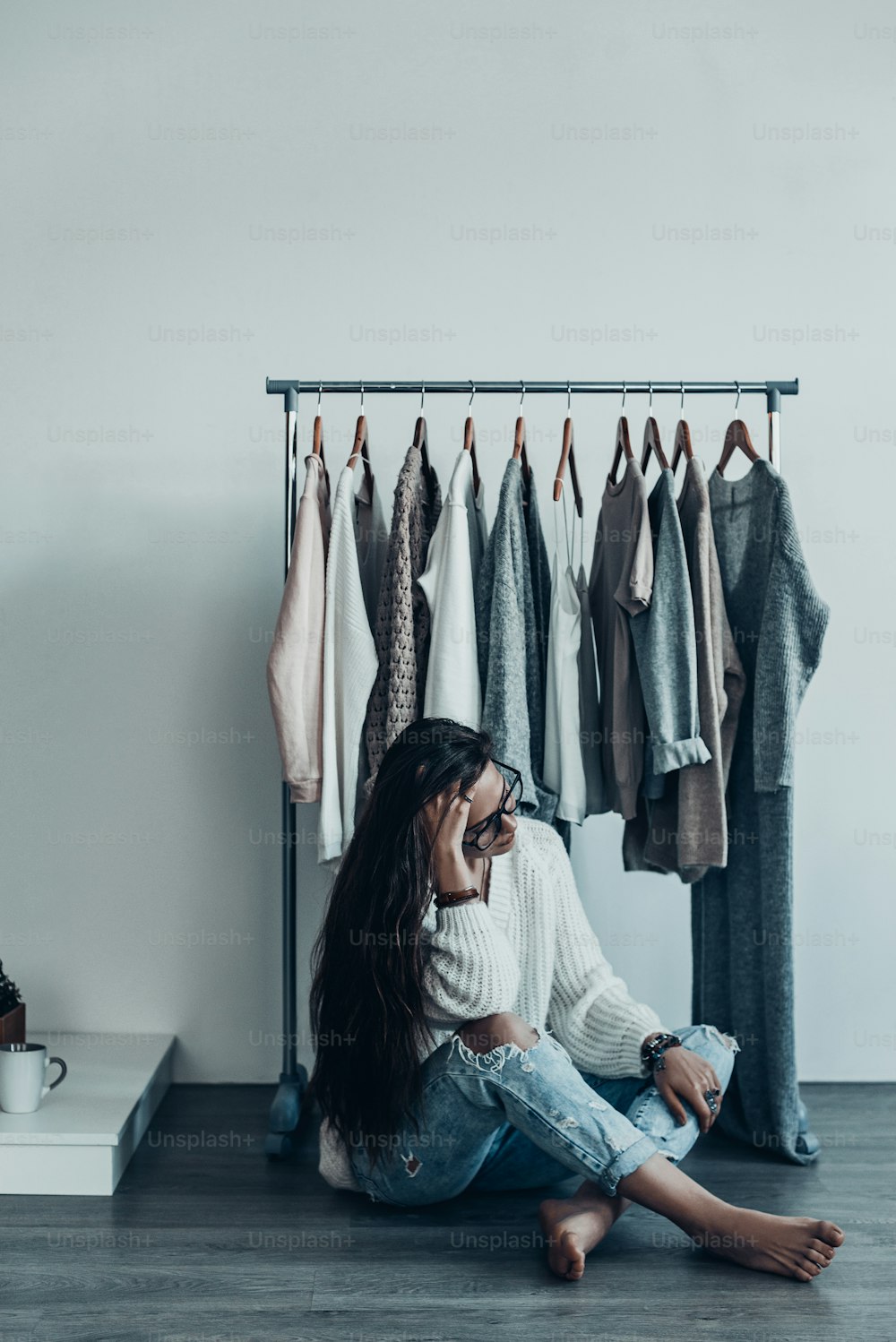 Thoughtful young woman in casual wear sitting on the floor at home and holding hand in hair near her clothes hanging on the racks