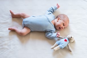 Cute newborn baby boy in blue striped onesie lying on bed, teddy bear toy with red heart next to him