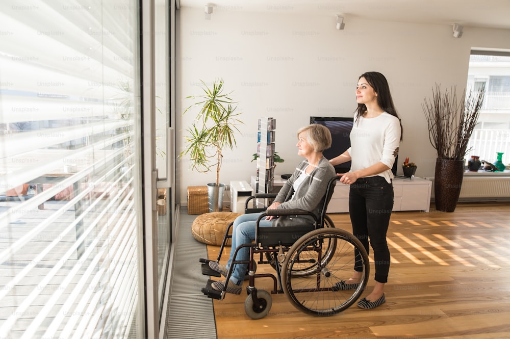 Une femme âgée handicapée en fauteuil roulant à la maison dans son salon, avec sa jeune fille qui s’occupe d’elle, regardant par la fenêtre.