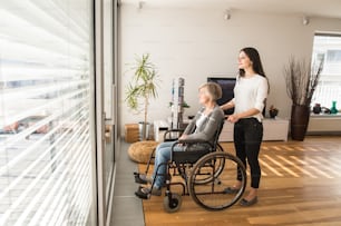 Behinderte ältere Frau im Rollstuhl zu Hause in ihrem Wohnzimmer, ihre kleine Tochter kümmert sich um sie und schaut aus dem Fenster.