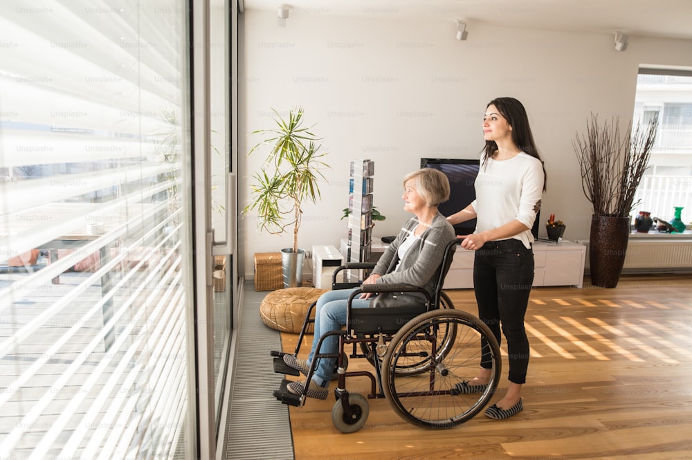 Disabled senior woman in wheelchair at home in her living room, with her young daughter caring for her, looking out the window.