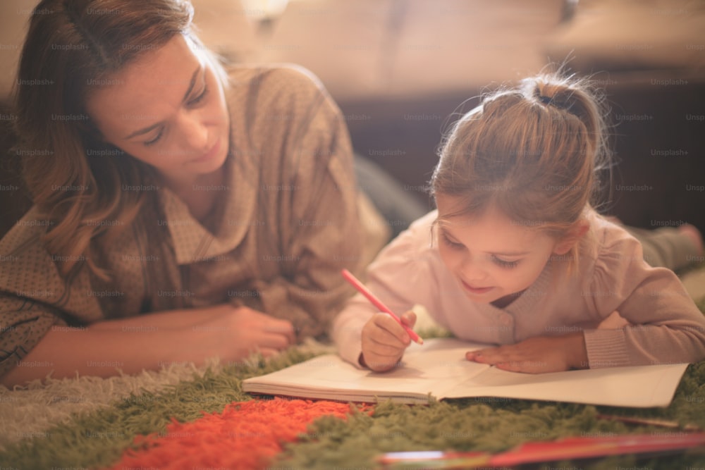 Beautiful blonde Caucasian girl lying on the floor with her mother and drawing.