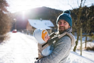Bel giovane padre con suo figlio fuori a fare una passeggiata, tenendolo nel marsupio. Natura invernale soleggiata.