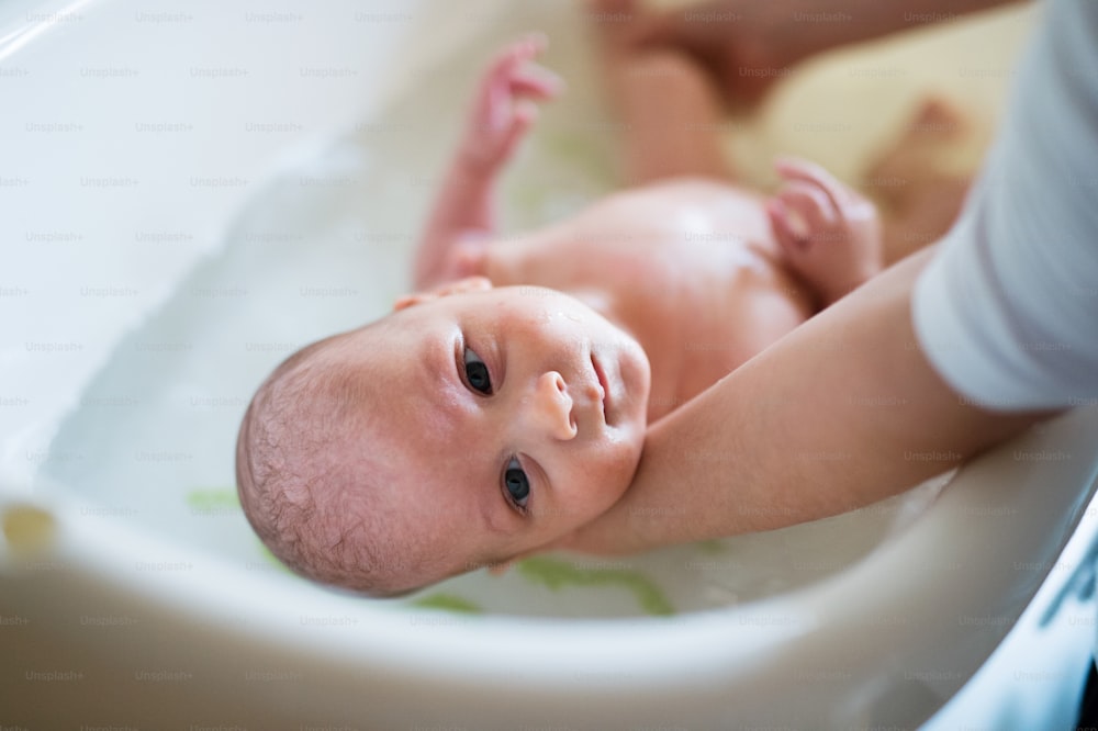 赤ん坊の息子を抱きかかえ、小さな白いプラスチックの風呂に入浴させている見分けのつかない母親。クローズアップ。