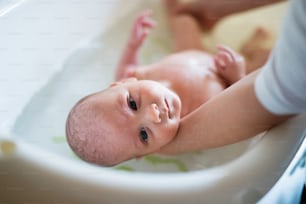 Unrecognizable mother holding her baby son, bathing him in small white plastic bath. Close up.