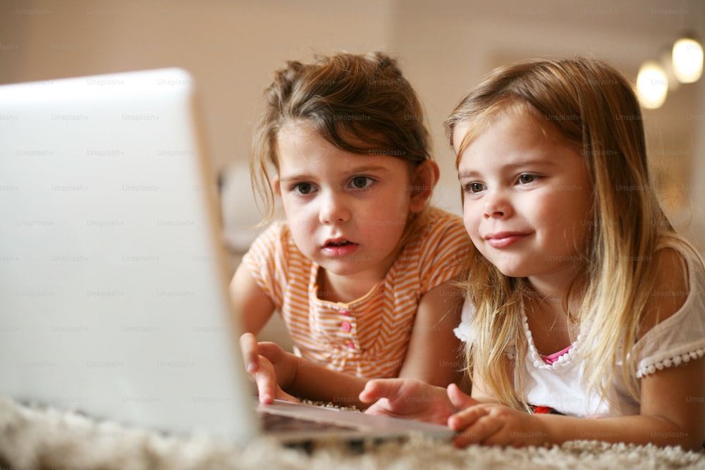 Two little sisters lying on the floor at living room and using laptop.