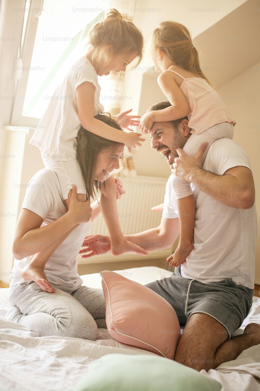 Family having funny pillow fight on bed. Parents spending free time with their daughters. Little girls sitting on parents shoulders.