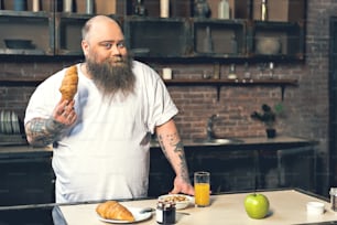 Eat only fresh food. Portrait of cheerful fat bearded man showing croissant while standing in kitchen. He is looking at camera and smiling. Copy space