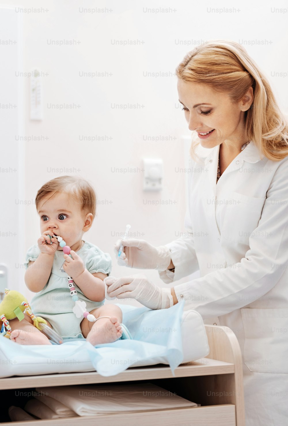 Wait a second. Charming baby girl looking forward playing with toy sitting near nurse