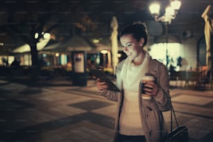 Chica usando tableta por la noche afuera y bebiendo café