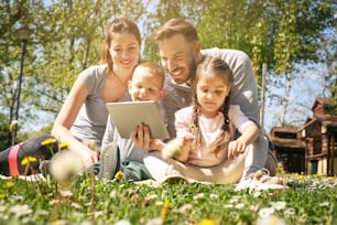 야외에서 태블릿을 사용하는 행복한 가족, 푸른 잔디에 앉아 있습니다.
