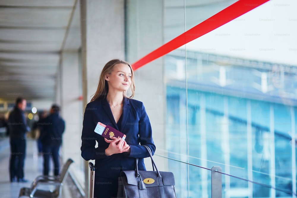 Young elegant business woman with hand luggage looking through the window in international airport terminal, waiting for her flight