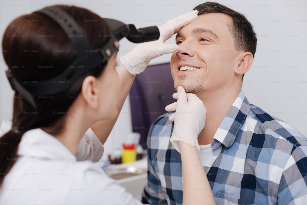 Time for work. Attentive otolaryngologist wearing ENT head light touching chin and forehead of her visitor sitting in semi position