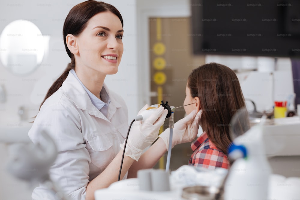 It will show us. Beautiful young female doctor wearing white smock keeping smile on her face while examining ear of little patient