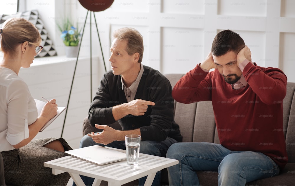 I do not want to hear you. Serious stubborn bearded man sitting on the sofa and pressing his hands to the ears while being unwilling to listen