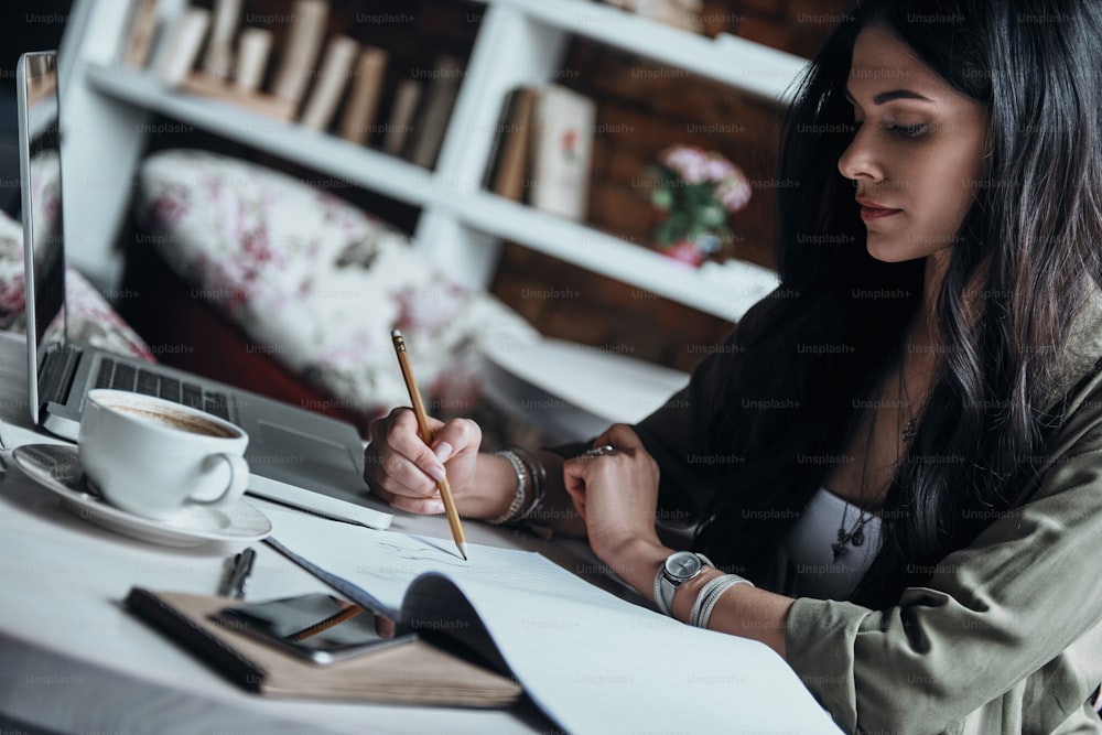 Nachdenkliche junge Frau, die etwas in ihr Notizbuch schreibt, während sie an ihrem Arbeitsplatz sitzt