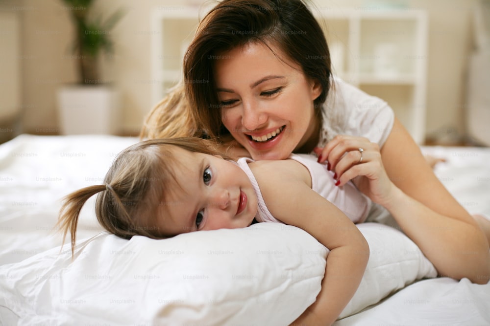 침대에서 딸과 놀고 있는 젊은 어머니. 어머니는 작고 귀여운 아기와 함께 자유 시간을 즐기고 있습니다.