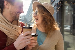 Love between us. Loving mature couple with coffee cups against city background. They are standing on the street and looking at each other with joy. Focus on cups